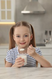 Photo of Cute little girl with tasty yogurt at white marble table in kitchen