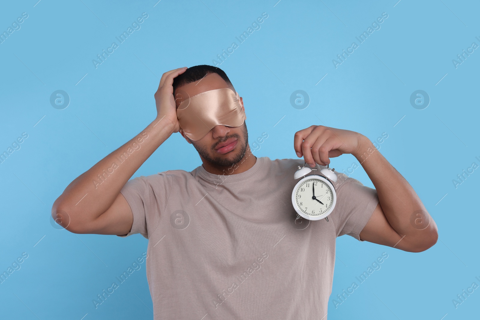 Photo of Tired man with sleep mask and alarm clock on light blue background. Insomnia problem