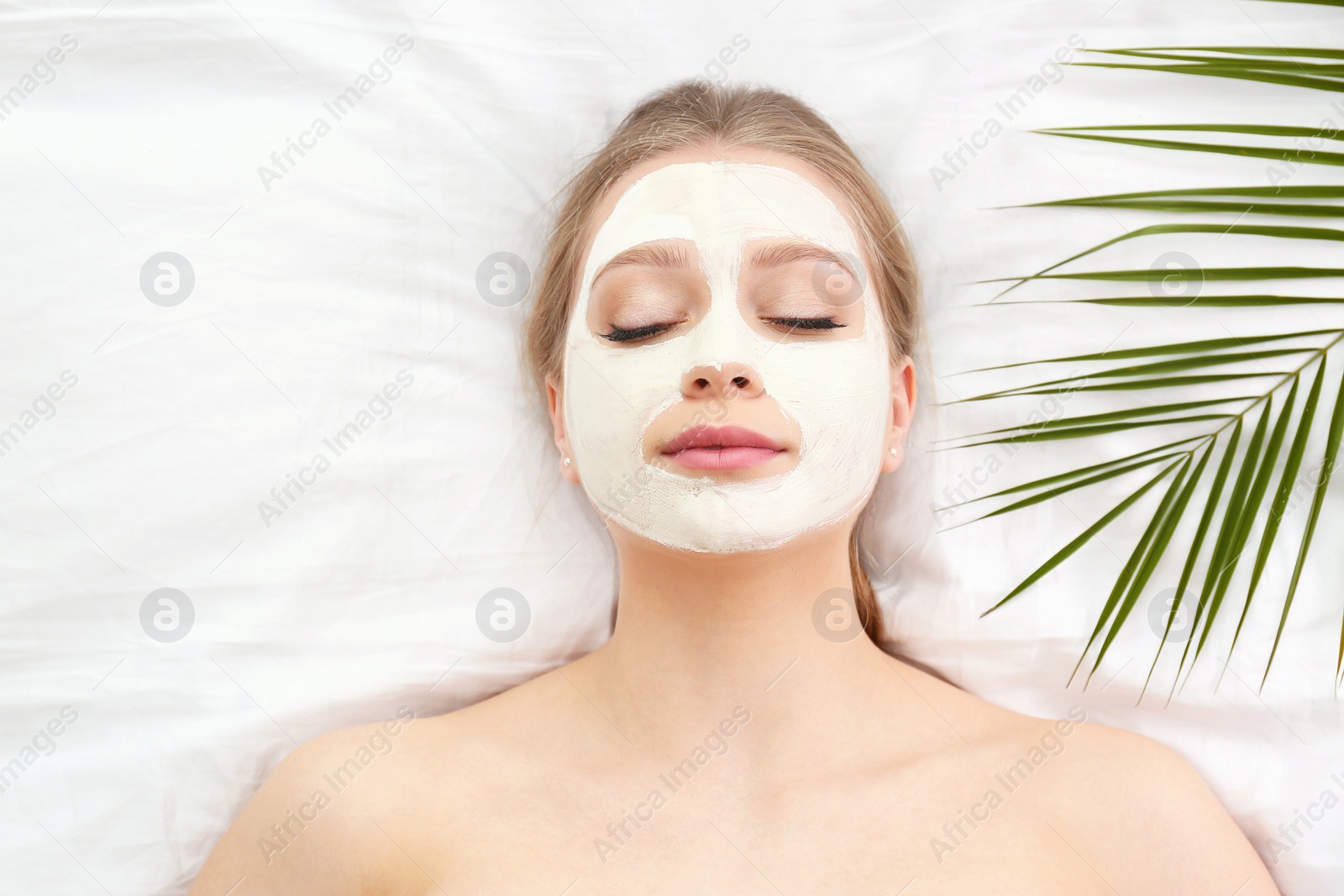 Photo of Beautiful woman with clay facial mask and tropical leaf on white fabric, above view