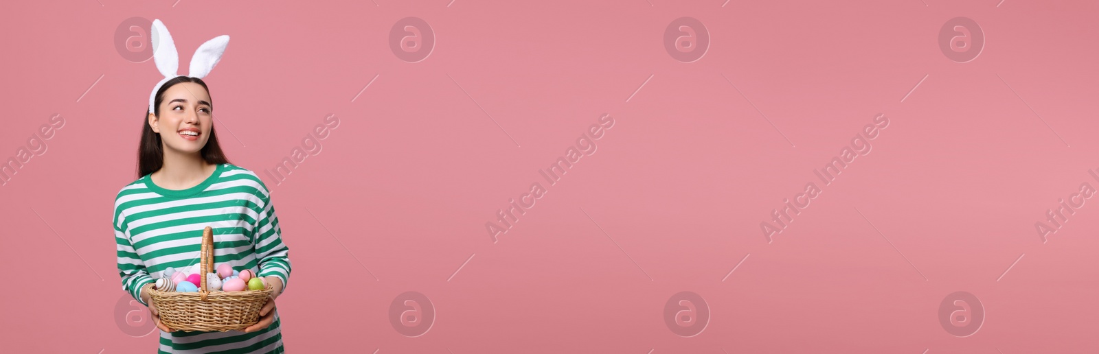 Image of Happy woman with bunny ears holding basket full of Easter eggs on pink background, space for text. Banner design