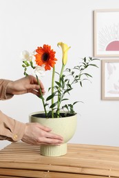 Photo of Stylish ikebana as house decor. Woman creating floral composition with fresh flowers at wooden table near white wall, closeup