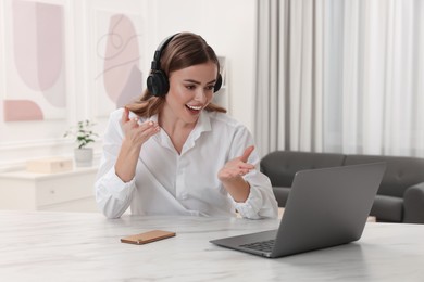 Happy woman with headphones having video chat via laptop at white table in room