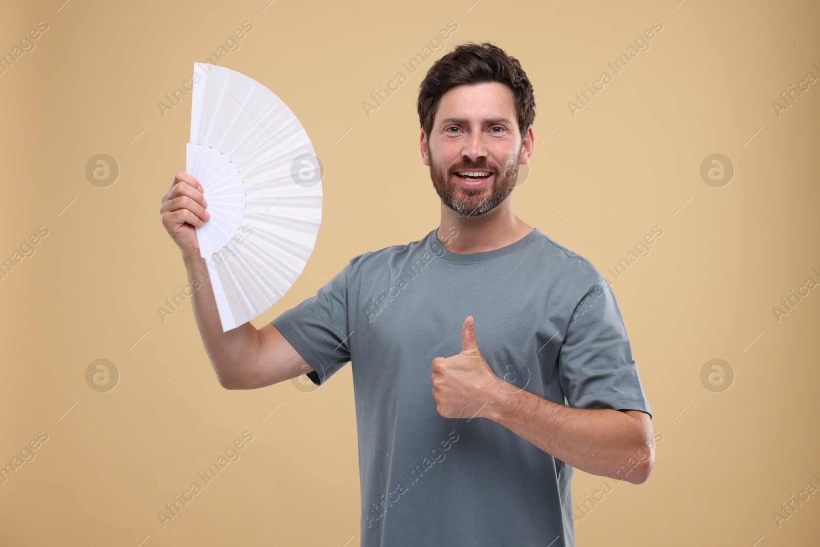 Photo of Happy man holding hand fan and showing thumb up on beige background