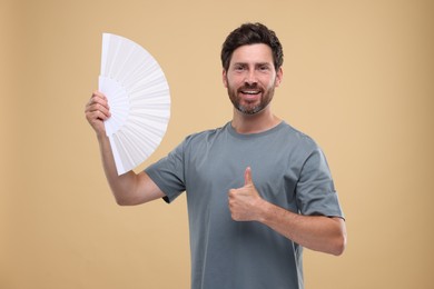 Photo of Happy man holding hand fan and showing thumb up on beige background