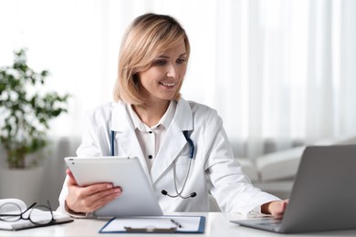 Photo of Smiling doctor with tablet and laptop having online consultation at table in office