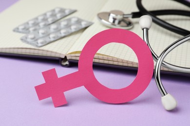 Photo of Female gender sign near open notebook, stethoscope and blisters of pills on violet background, closeup. Women's health concept