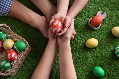 Photo of Father, mother and their child holding painted Easter egg on green grass, top view