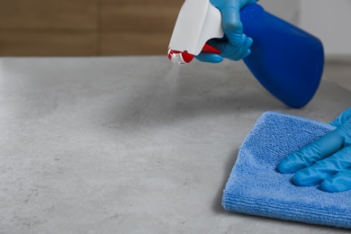 Photo of Woman in gloves cleaning light grey stone table with rag and detergent indoors, closeup. Space for text