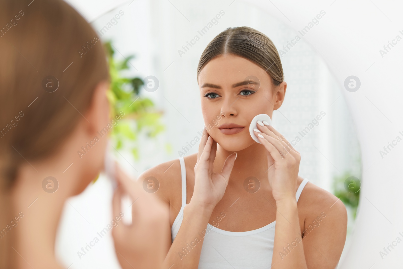 Photo of Beautiful woman removing makeup with cotton pad near mirror indoors