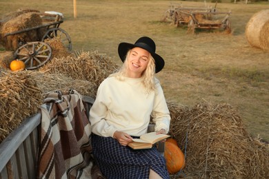 Photo of Beautiful woman with book sitting on wooden bench near hay bales outdoors. Autumn season