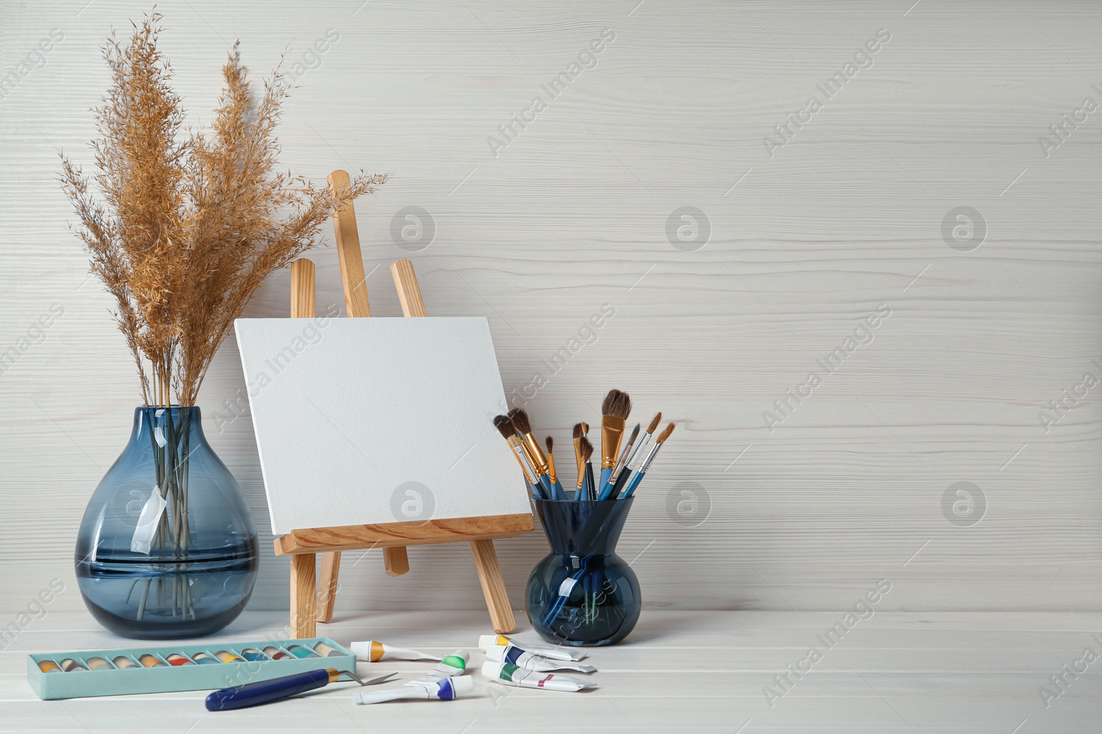 Photo of Easel with blank canvas, paints and brushes on white wooden table. Space for text