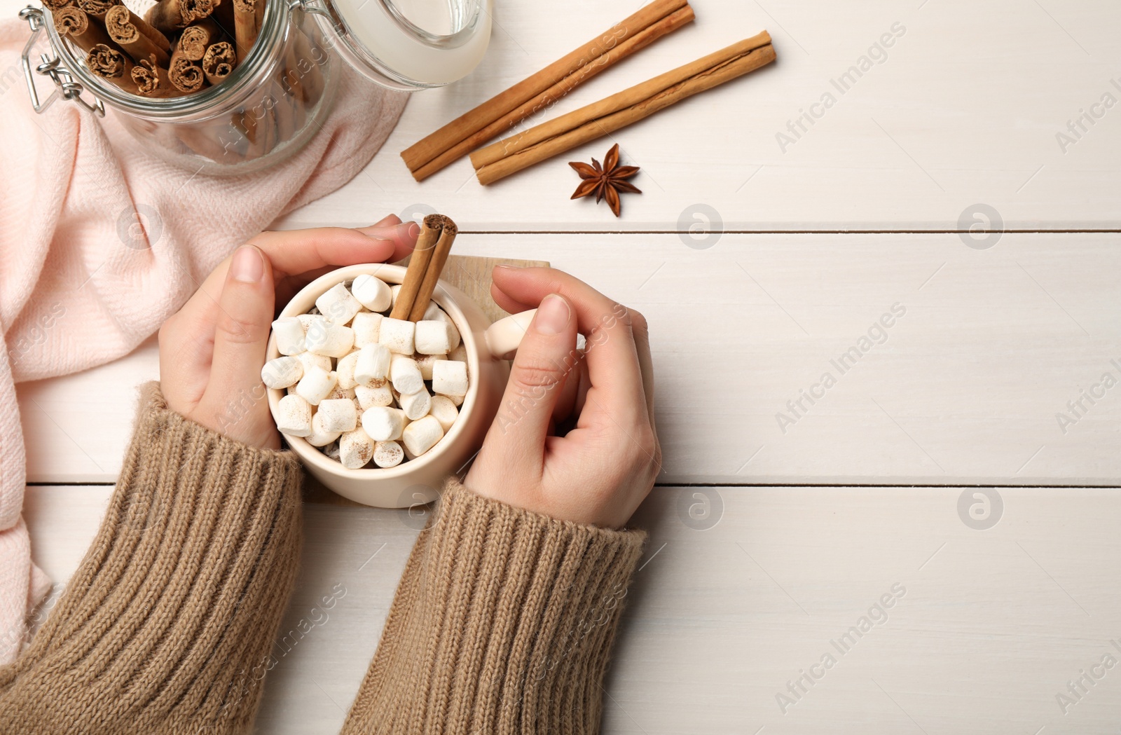 Photo of Woman holding cup of hot drink with marshmallows and aromatic cinnamon at white wooden table, top view. Space for text