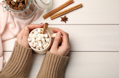 Photo of Woman holding cup of hot drink with marshmallows and aromatic cinnamon at white wooden table, top view. Space for text