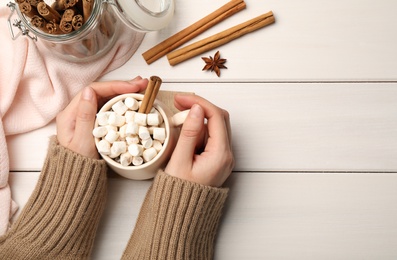 Woman holding cup of hot drink with marshmallows and aromatic cinnamon at white wooden table, top view. Space for text