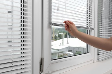 Photo of Woman opening horizontal blinds on window indoors, closeup