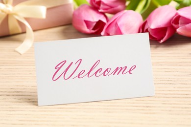 Image of Welcome card, gift box and beautiful pink tulips on wooden table, closeup