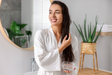 Photo of Young woman holding bowl of aloe hair mask in bathroom