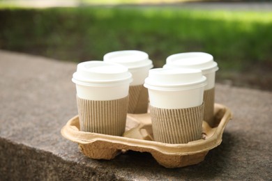 Photo of Takeaway paper coffee cups with plastic lids and sleeves in cardboard holder outdoors