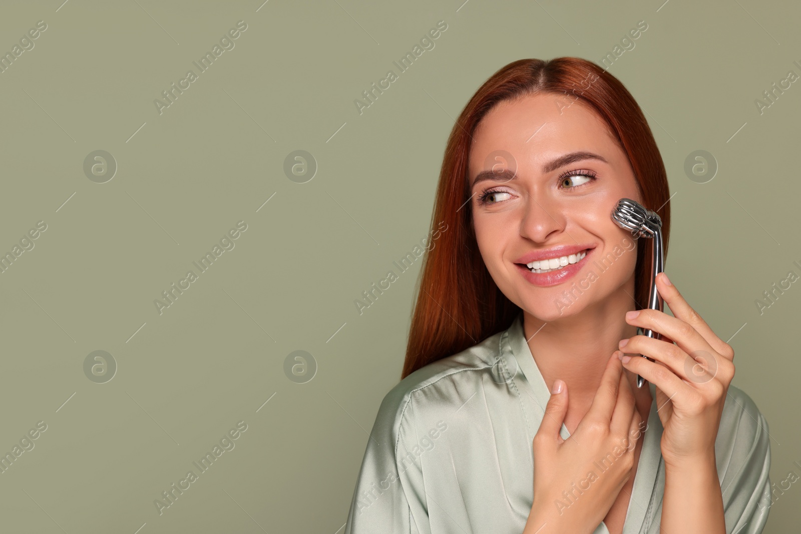 Photo of Young woman massaging her face with metal roller on green background, space for text