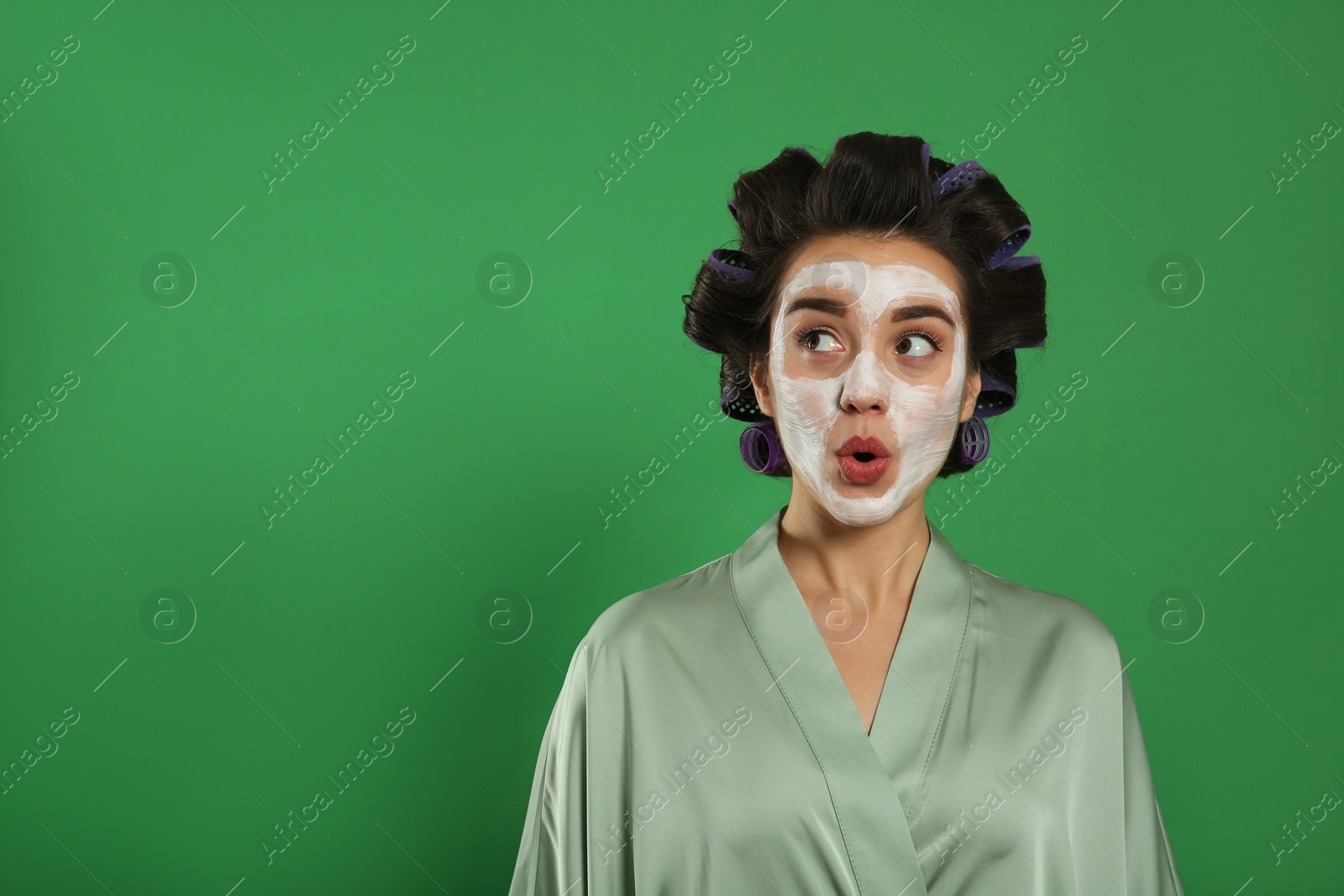 Photo of Emotional young woman in silk bathrobe with hair curlers and facial mask on green background, space for text