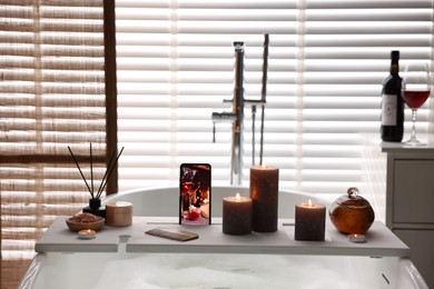 Photo of White wooden tray with smartphone, burning candles and beauty products on bathtub in bathroom