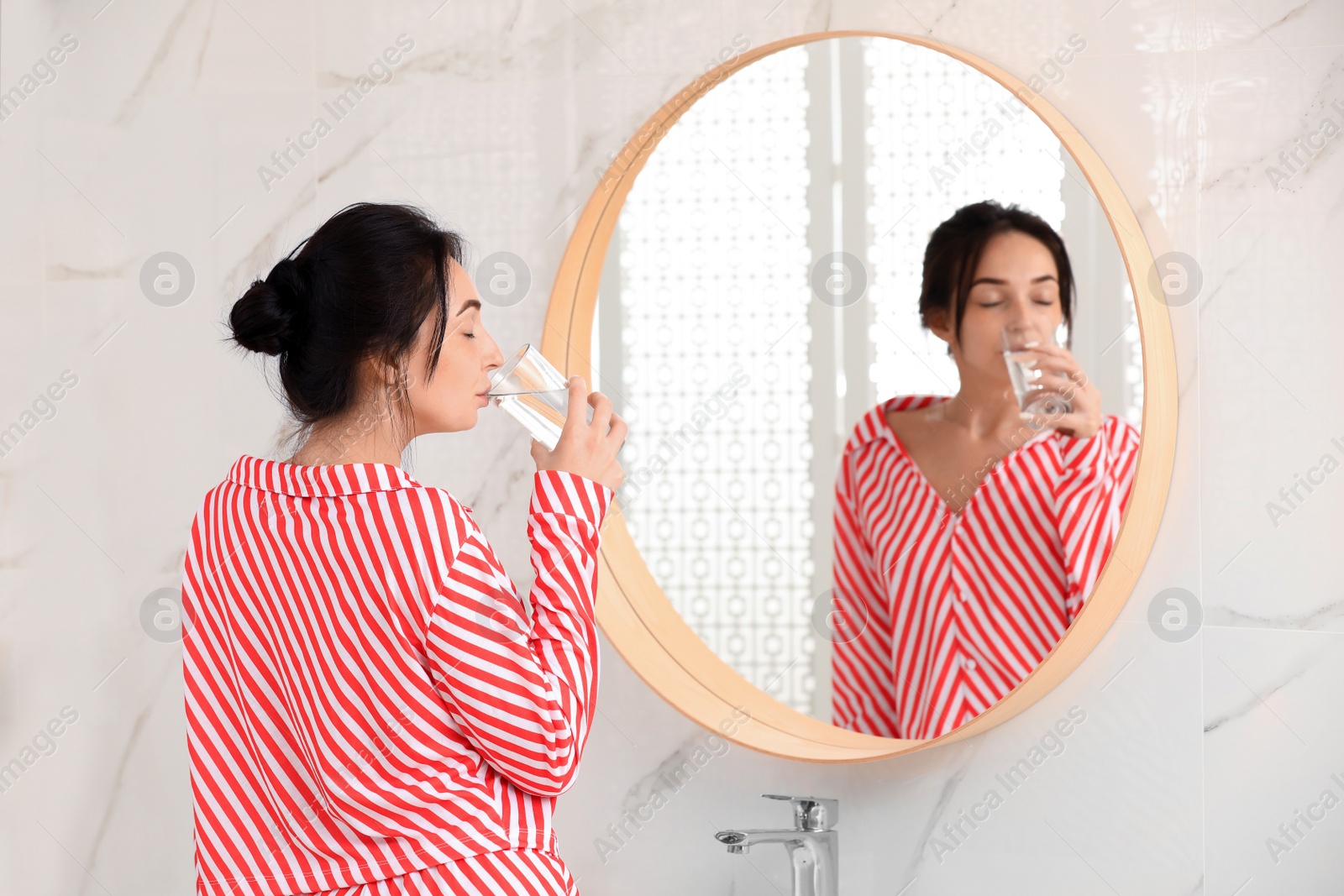 Photo of Young woman drinking water near mirror in bathroom