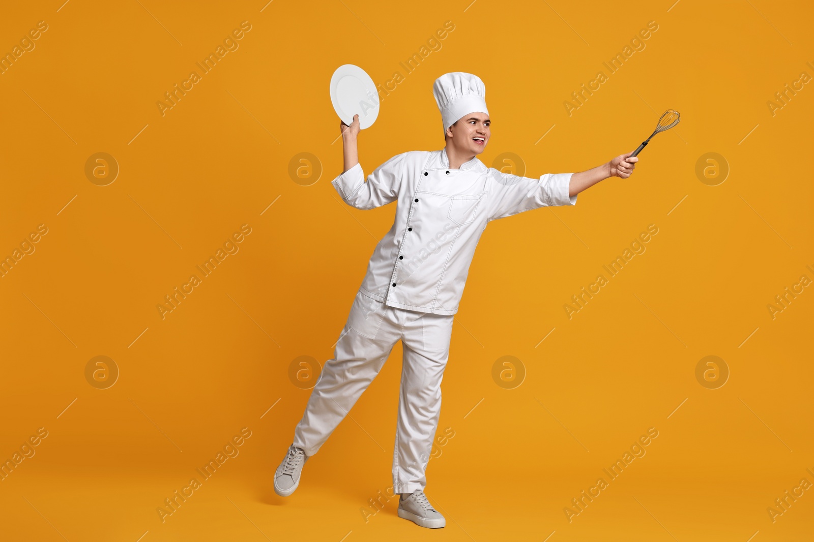 Photo of Portrait of happy confectioner in uniform holding plate and whisk on orange background