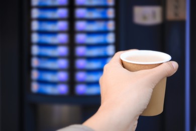 Man holding paper cup with hot drink near coffee vending machine, closeup. Space for text
