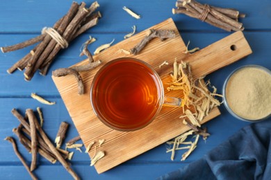 Aromatic licorice tea in cup, dried sticks of licorice root and powder on blue wooden table, flat lay