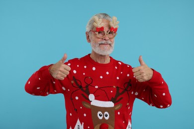 Photo of Senior man in Christmas sweater and funny glasses showing thumbs up on light blue background