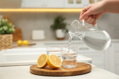 Photo of Woman pouring water from jug into glass at white table in kitchen, closeup