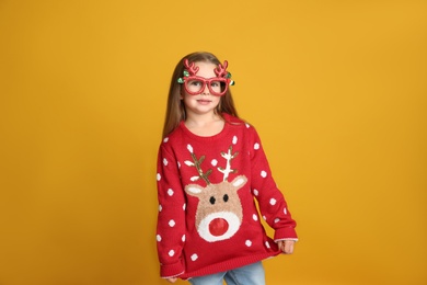 Cute little girl in Christmas sweater and party glasses on yellow background