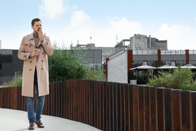 Photo of Sleepy man with cup of coffee yawning outdoors