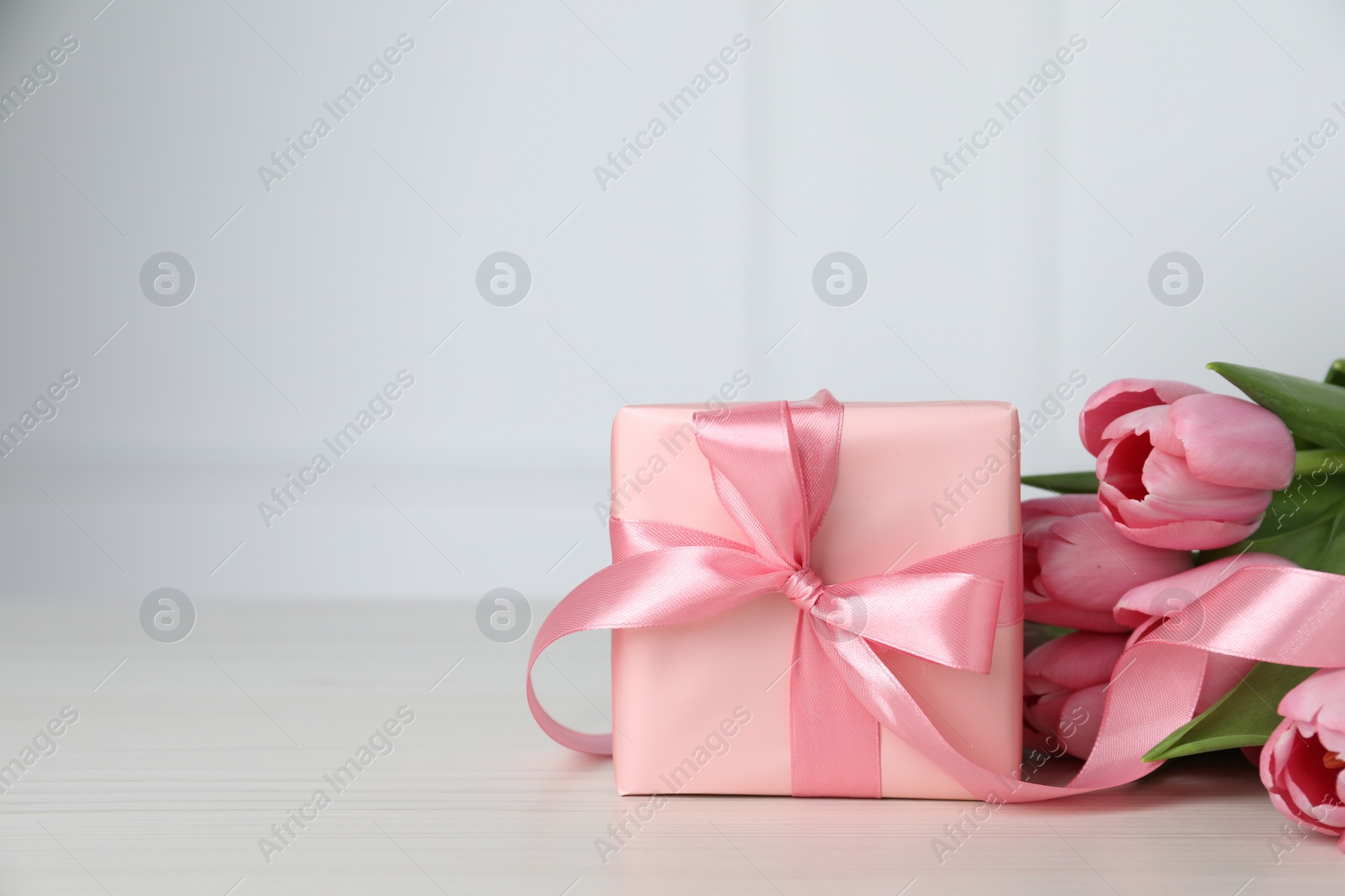 Photo of Beautiful gift box with bow and pink tulip flowers on light table, space for text