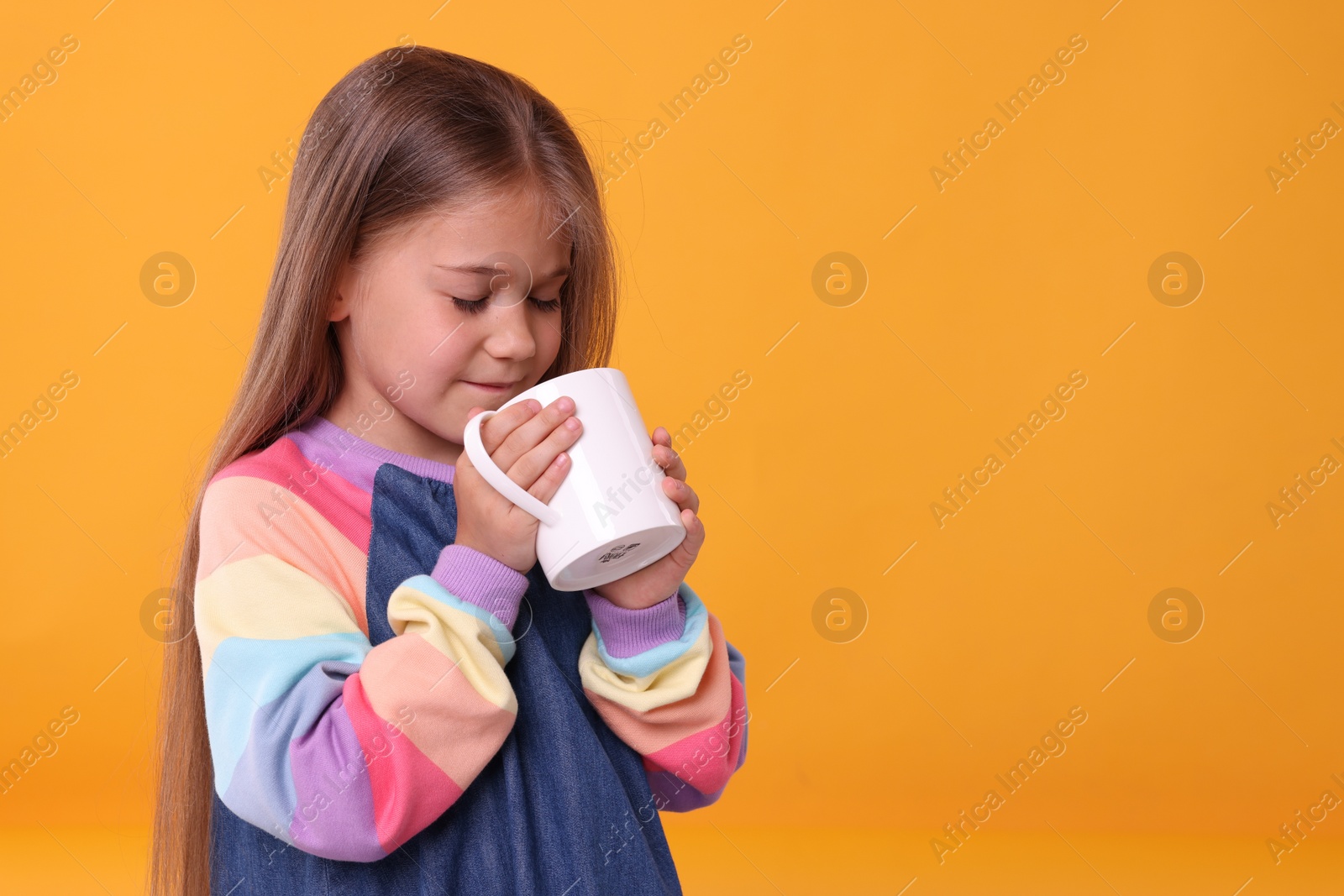 Photo of Cute girl drinking beverage from white ceramic mug on orange background, space for text