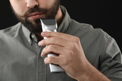 Handsome young man trimming beard on black background, closeup