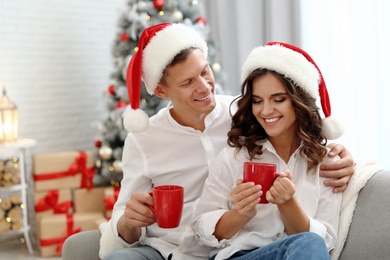 Happy couple in Santa hats with cups of hot drink near Christmas tree at home