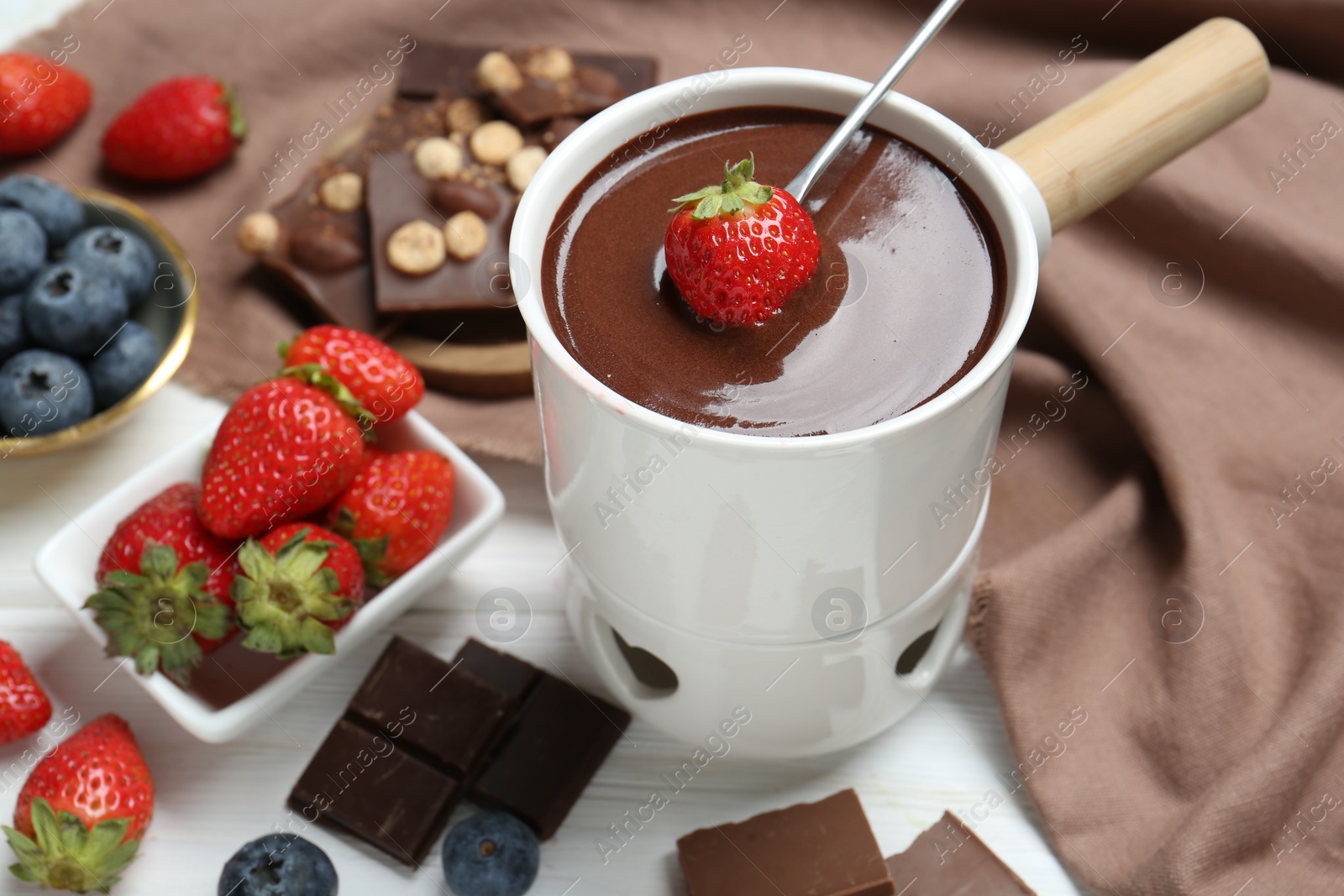 Photo of Dipping fresh strawberry in fondue pot with melted chocolate at white wooden table