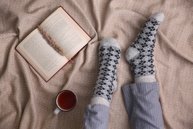 Woman with cup of hot winter drink and book on warm plaid, top view. Cozy season