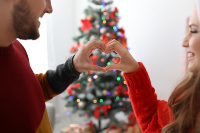 Photo of Young couple putting hands in shape of heart together near Christmas tree at home