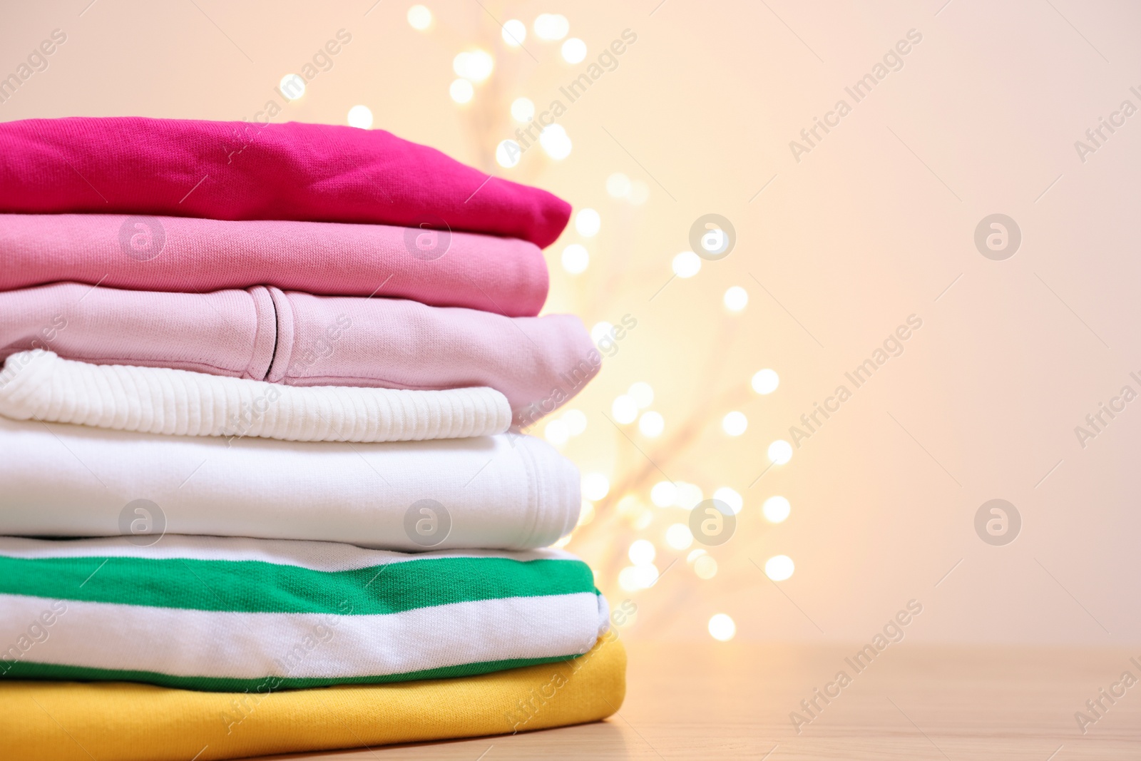 Stack of folded clothes on wooden table against beige background with blurred lights, space for text Photo of Stack of folded clothes on wooden table against beige background with blurred lights, space for text