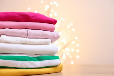 Photo of Stack of folded clothes on wooden table against beige background with blurred lights, space for text