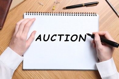 Image of Woman writing word Auction in notebook at wooden table, top view