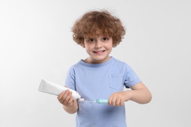 Photo of Cute little boy squeezing toothpaste from tube onto electric toothbrush on white background