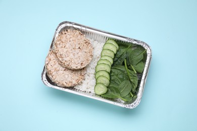 Container with rice, puffed cakes. fresh spinach and cut cucumbers on light blue background, top view