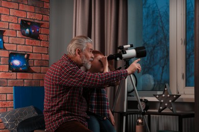 Photo of Little boy with his grandfather looking at stars through telescope in room