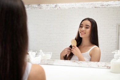 Photo of Beautiful young woman with hair brush looking into mirror in bathroom