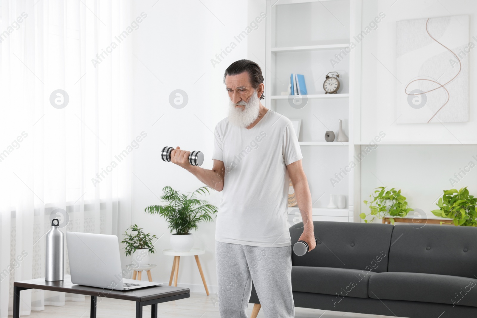 Photo of Senior man exercising with dumbbells while watching online tutorial at home. Sports equipment