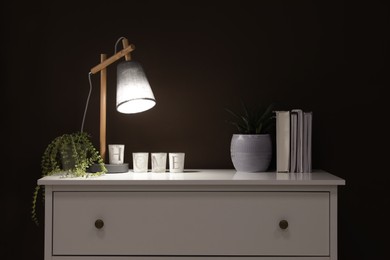 Photo of Stylish lamp, books and green plants on white chest of drawers near brown wall indoors. Interior design