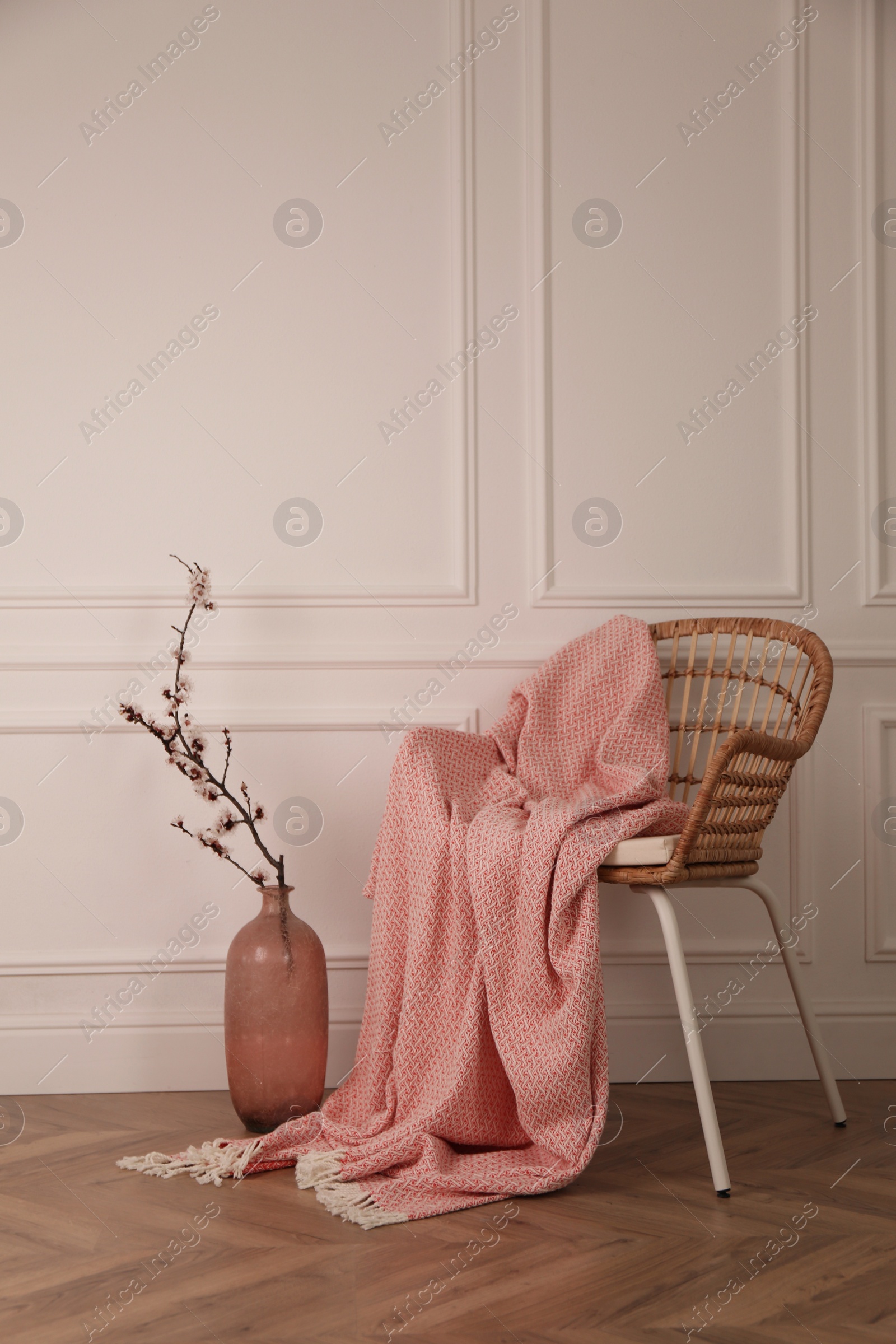 Photo of Flowering tree twig in glass vase on floor near chair with blanket at white wall